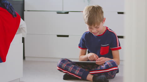 boy at home in bedroom wearing pyjamas playing on digital tablet sitting on floor