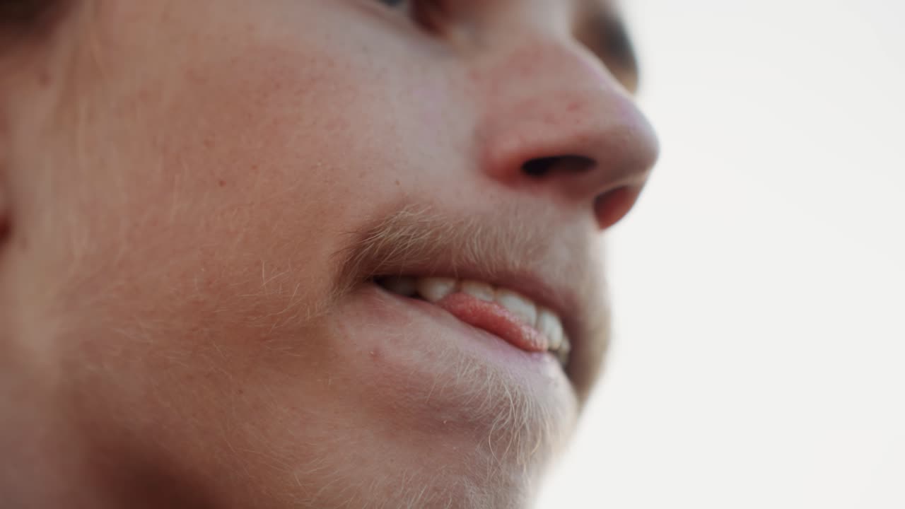 Adolescente bianco si morde il labbro inferiore, espressione pensierosa e ansiosa durante un momento di quiete, mascella lentigginosa con leggera barba incolta, primo piano di bocca e mento, umore introspettivo, gesto sottile delle dita, naturale e delicato.