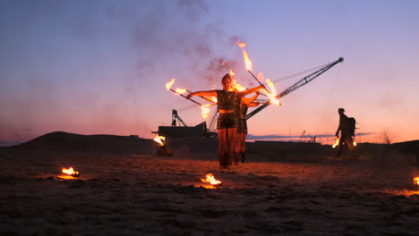 a group of men and woman fire show at night on the sand against the background of fire and tower cranes.