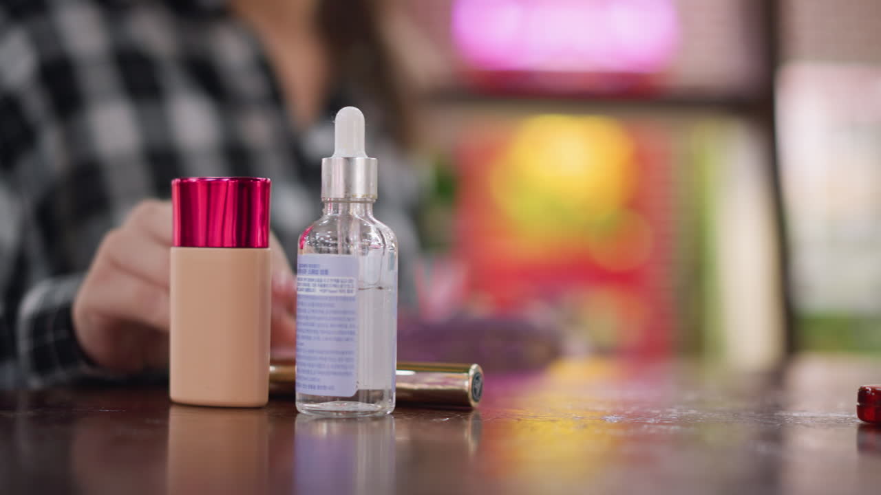 나무 테이블 위에 메이크업 도구를 놓고 있는 카레드 셔츠를 입은 여자의 흐릿한 시선, 전면에 드롭퍼가 있는 유리 세럼 병에 초점을 맞추고, 부드러운 조명과 따뜻한 배경 톤으로 아름다움 루틴 설정