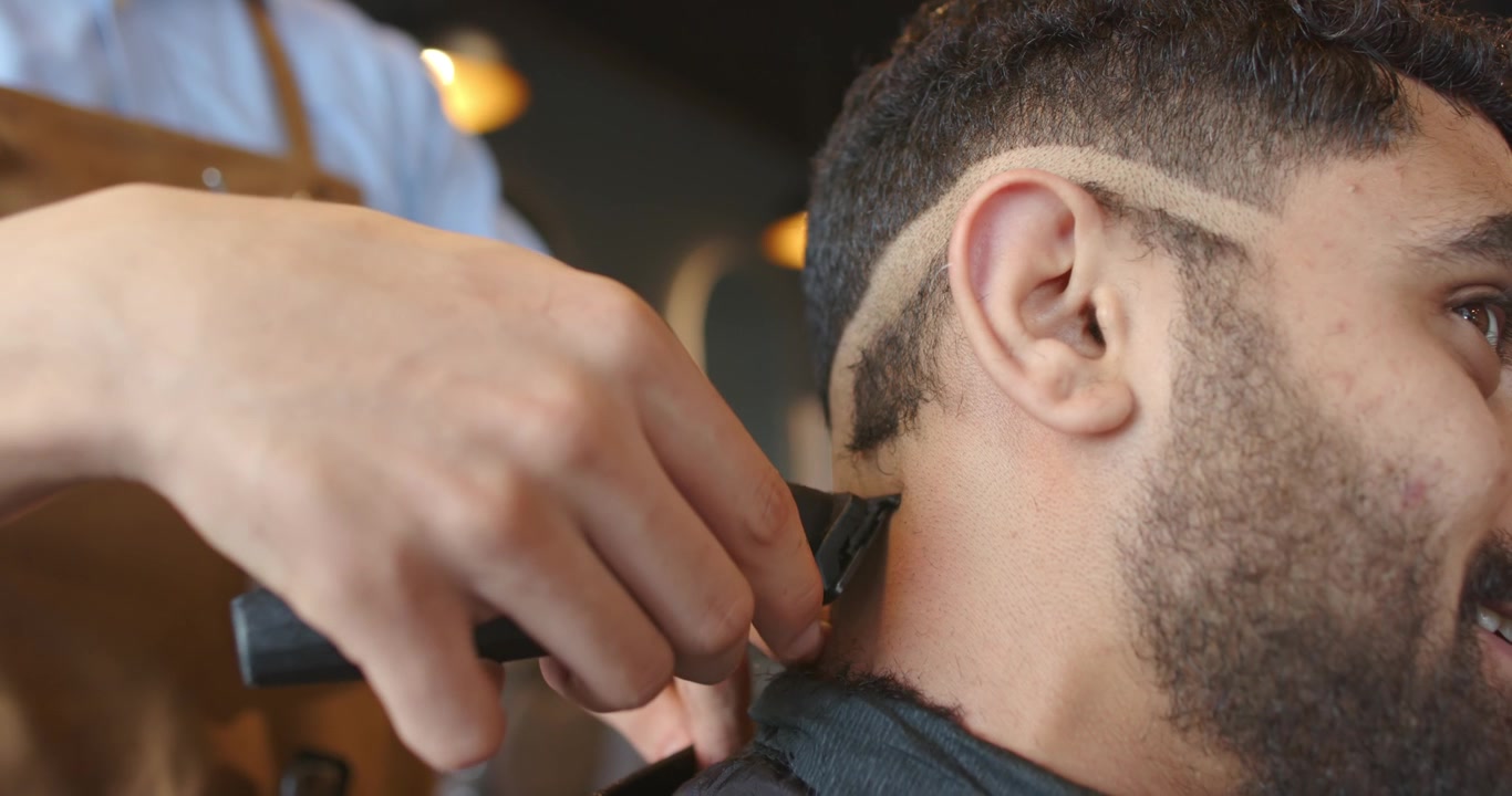 Barbeiro com avental de couro posicionando a máquina de cortar cabelo no pescoço de um homem na barbearia, aparando, refinando o degradê.