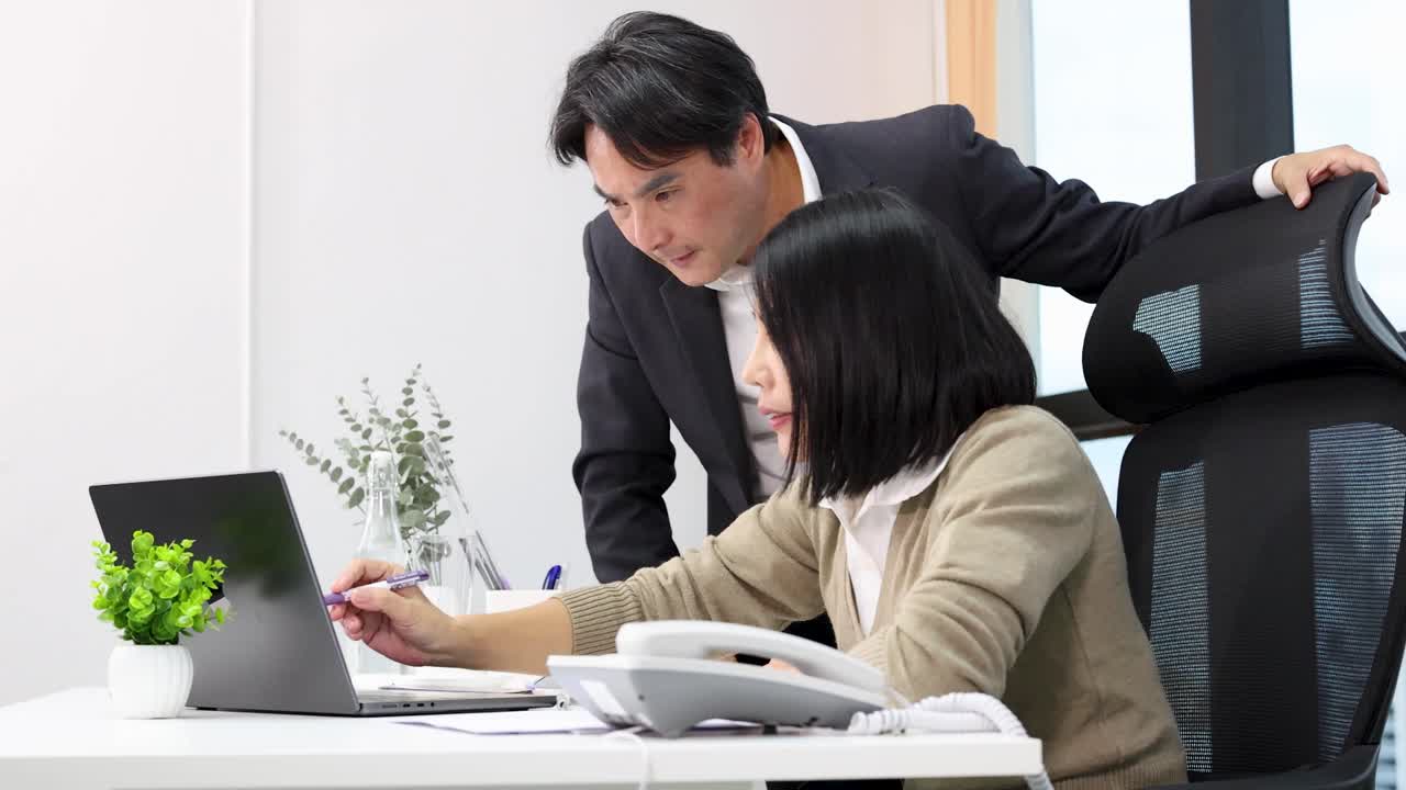 明るいオフィスで、アジア系のマネージャーと従業員がノートパソコンの画面を見ながら仕事について話し合っています。