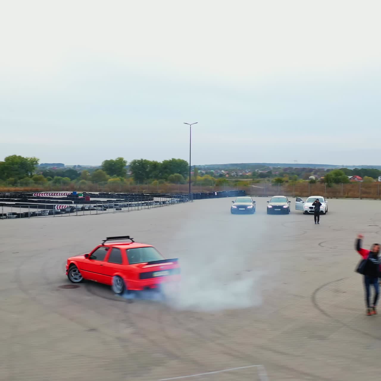Red car riding circles drifting at the square. Bunch of people dancing hip hop at backdrop. High angle view