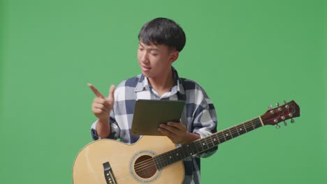 adolescente asiático con guitarra sosteniendo una tableta y apuntando hacia un lado mientras está de pie en el fondo de la pantalla verde