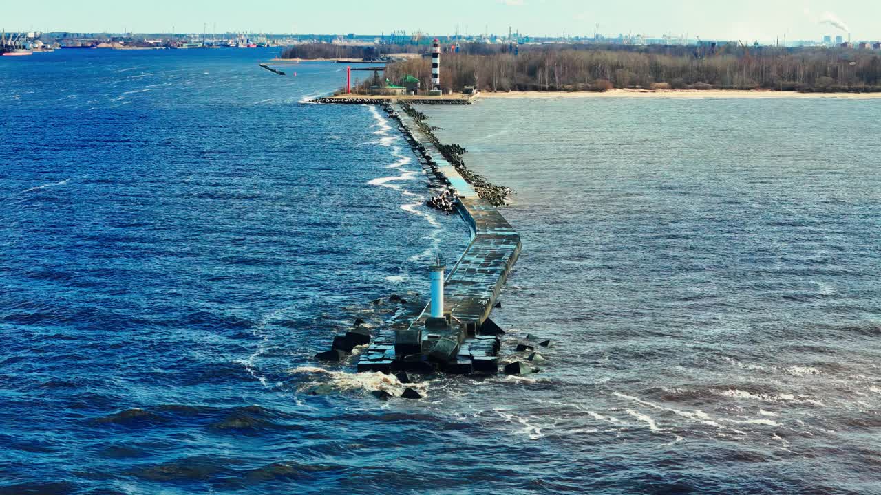 A long stone jetty extends into bright blue Baltic Sea, ending at a tall black and white lighthouse dividing choppy waves from muted estuary waters and sandy coastline.