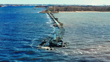 a long stone jetty extends into bright blue baltic sea, ending at a tall black and white lighthouse dividing choppy waves from muted estuary waters and sandy coastline.