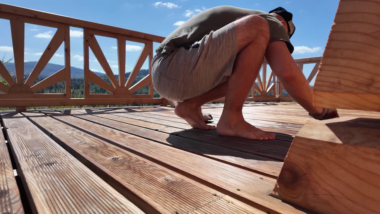 Man in hat oils deck boards in summer sun. Decorative railing, gloves, brush, and mountain horizon highlight home renovation and outdoor wood preservation