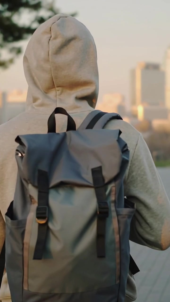 Vertical video: Turning, hooded walker fixing backpack strap while walking on plaza to city skyline