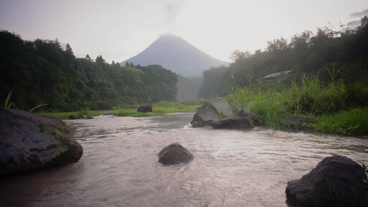 Mountain water stream flowing between rocks with volcano that emits smoke - beautiful nature footage in jungle