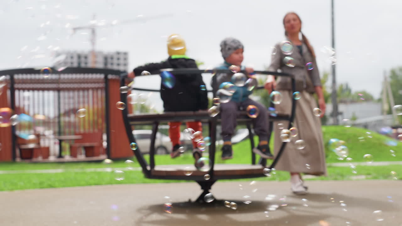 Colorful bubbles drift across foreground as mother rotates child on merry go round, soft focus background with playground structures, green lawn and overcast sky, joyful motion, gentle spin