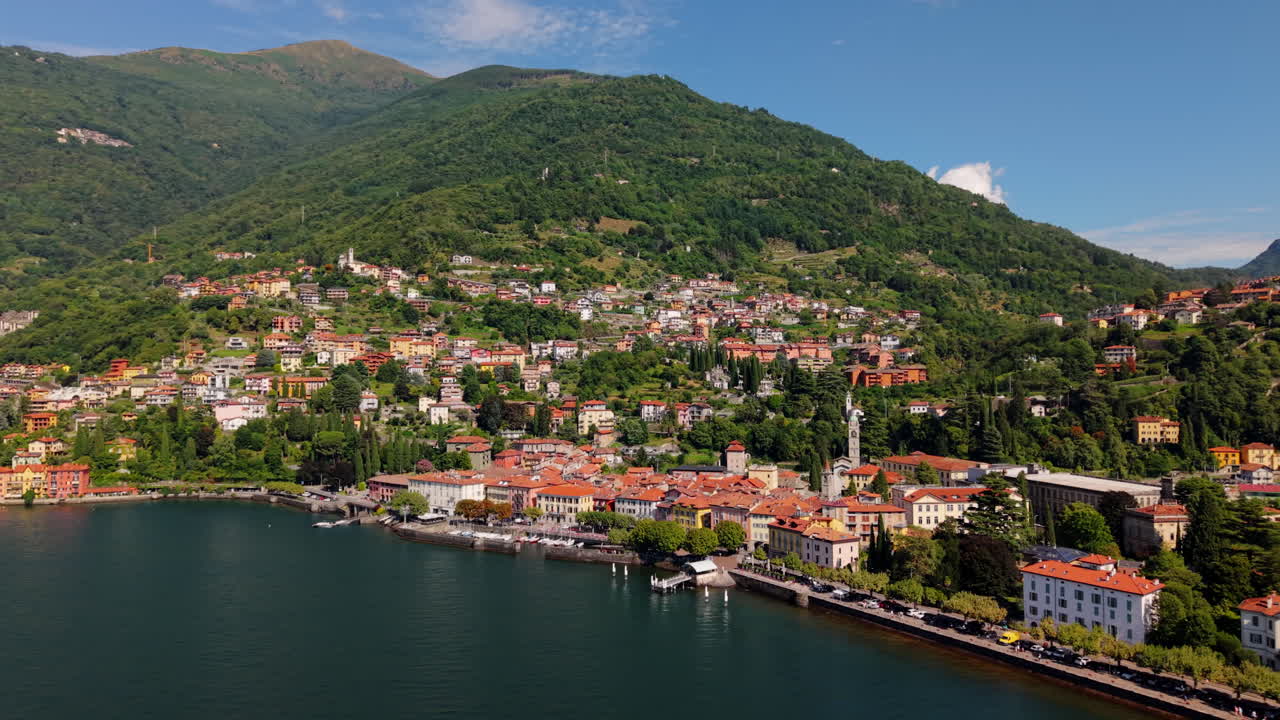 Drone flies forward in a slight arc above Lake Como, revealing the waterfront town, red-roofed houses, and green mountains in clear daylight