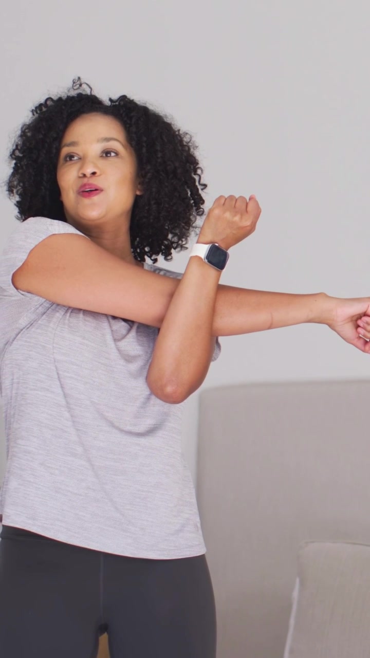 Vertical video: Exhaling woman in tee pulling arm over chest to stretch at home checking smartwatch