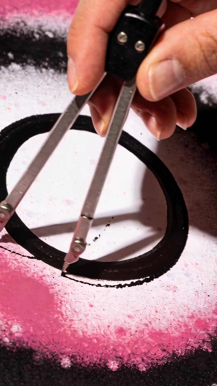 Vertical video: Placing nib, hand rotating compass on paper over pink spray refining black circle