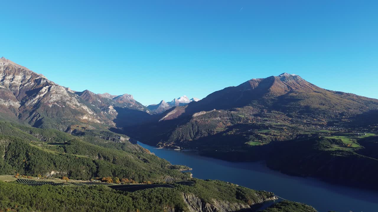 Drone captures the deep blue waters of Lake Serre Ponçon surrounded by rugged alpine peaks and autumn forests. Clear skies and November sunlight highlight the dramatic landscape in the Hautes Alpes