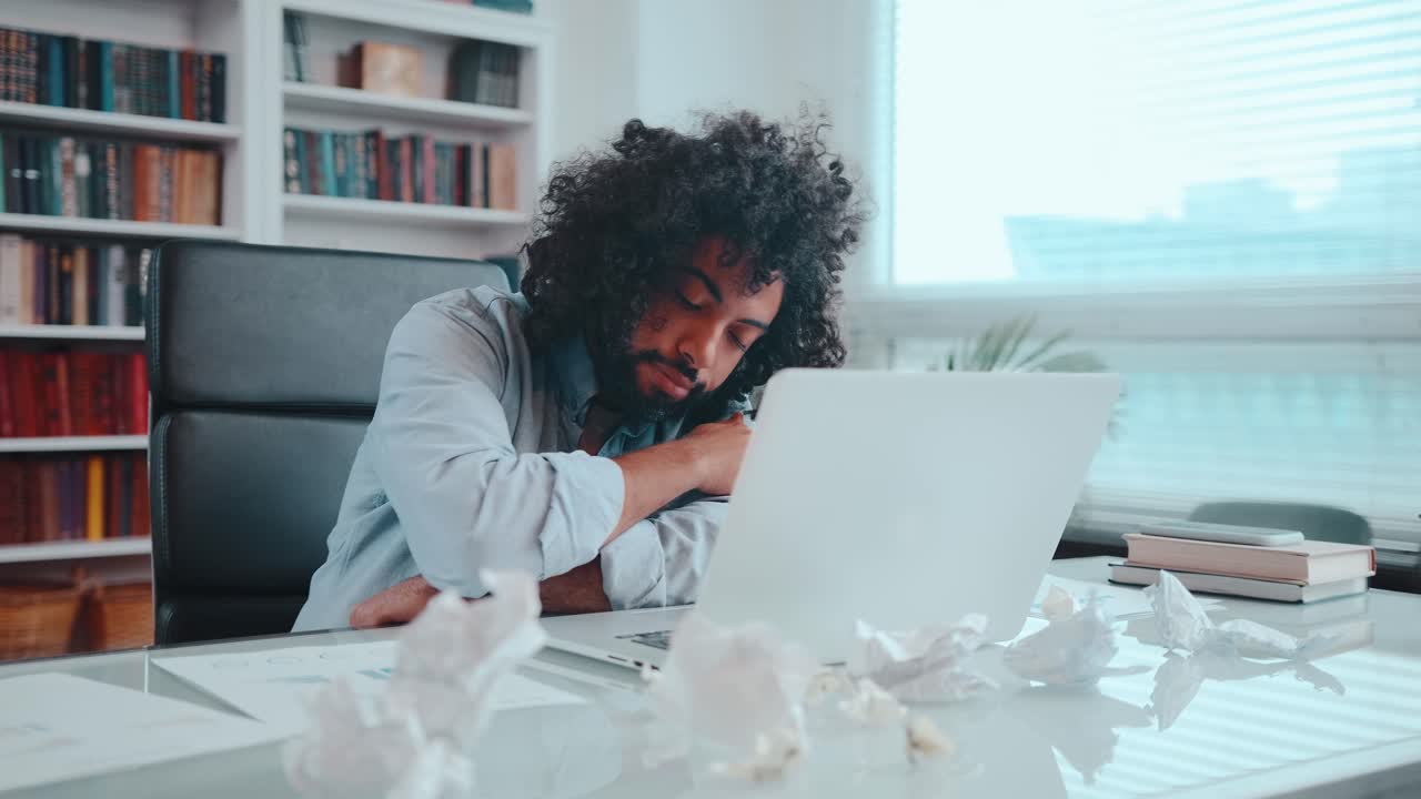 Young arabian man yawns and rests head on table next to laptop and falls asleep