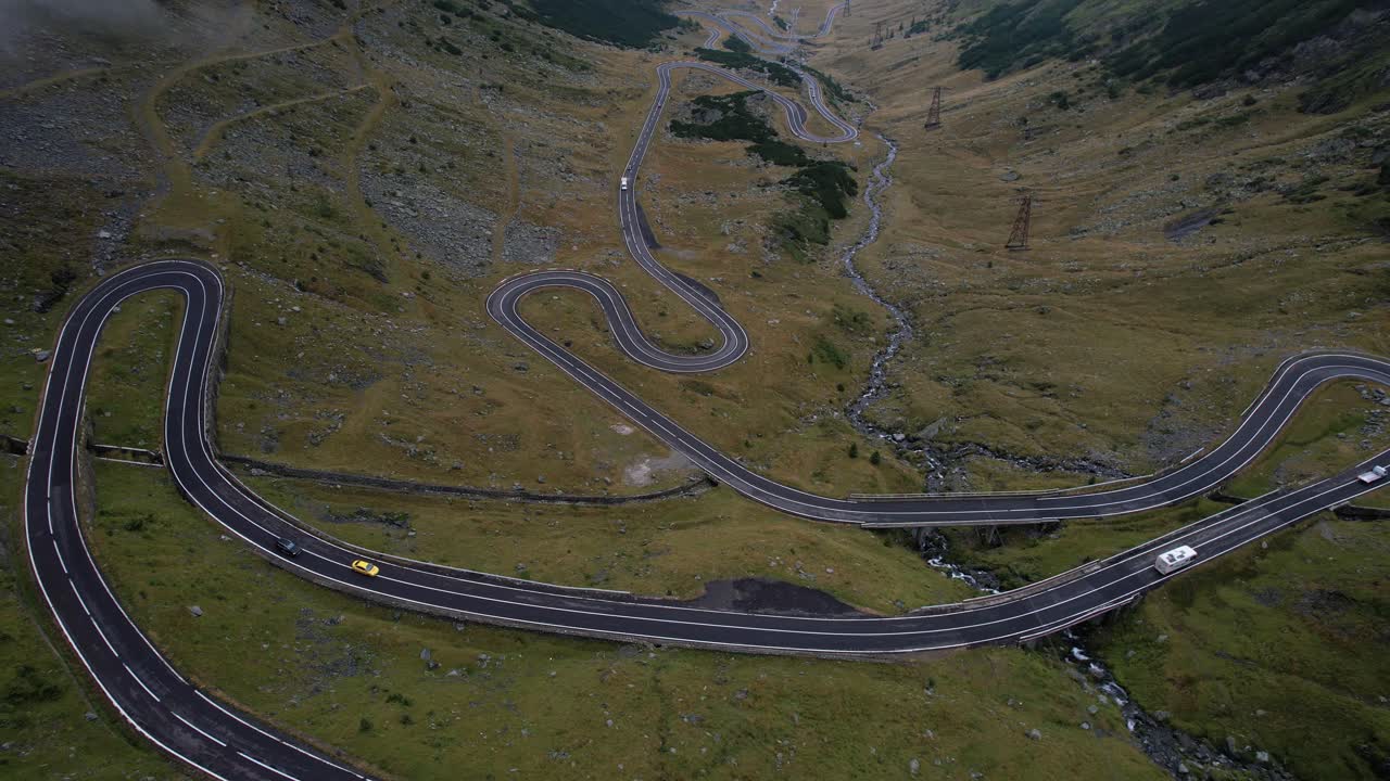 A cinematic aerial sliding reveal shot of the Transfagarasan road, moving from right to left, rising to unveil the lush valley, dramatic mountains, and soft clouds drifting across the landscape.