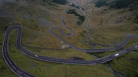 a cinematic aerial sliding reveal shot of the transfagarasan road, moving from right to left, rising to unveil the lush valley, dramatic mountains, and soft clouds drifting across the landscape.