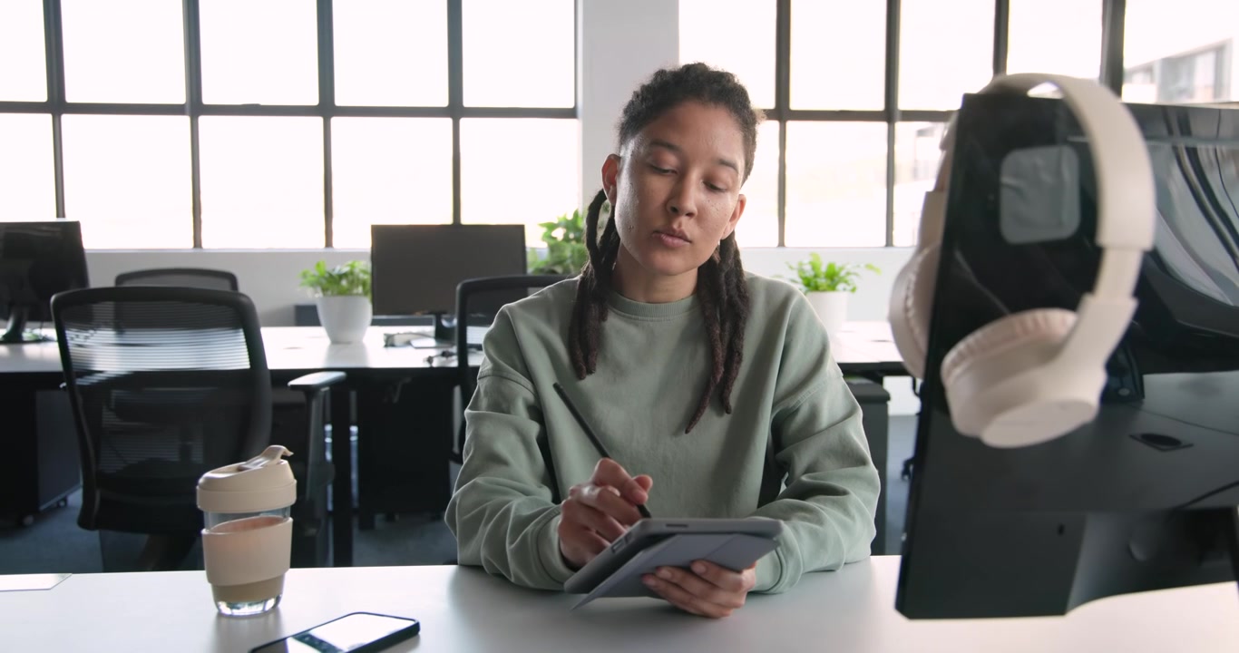 Non-binary African designer holding tablet and sketching with stylus for work at desk, phone cup