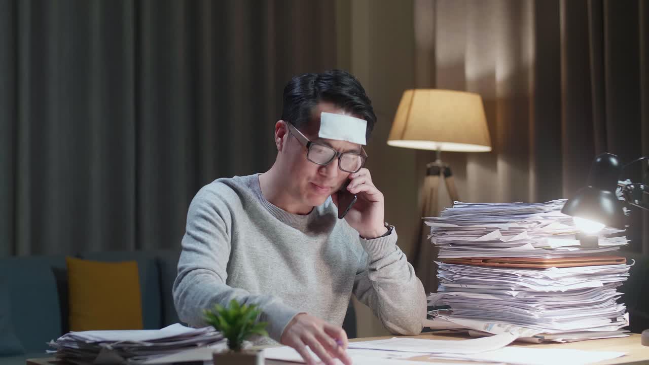 Sick Asian Man Talking On Phone And Taking Note While Working Hard With Documents At Home