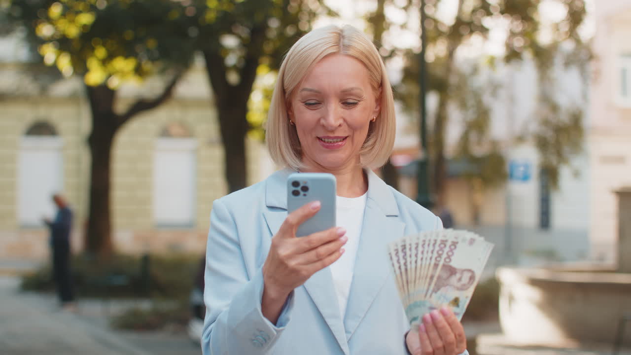 Caucasian mature businesswoman on city street showing earnings cash money with smartphone in hands