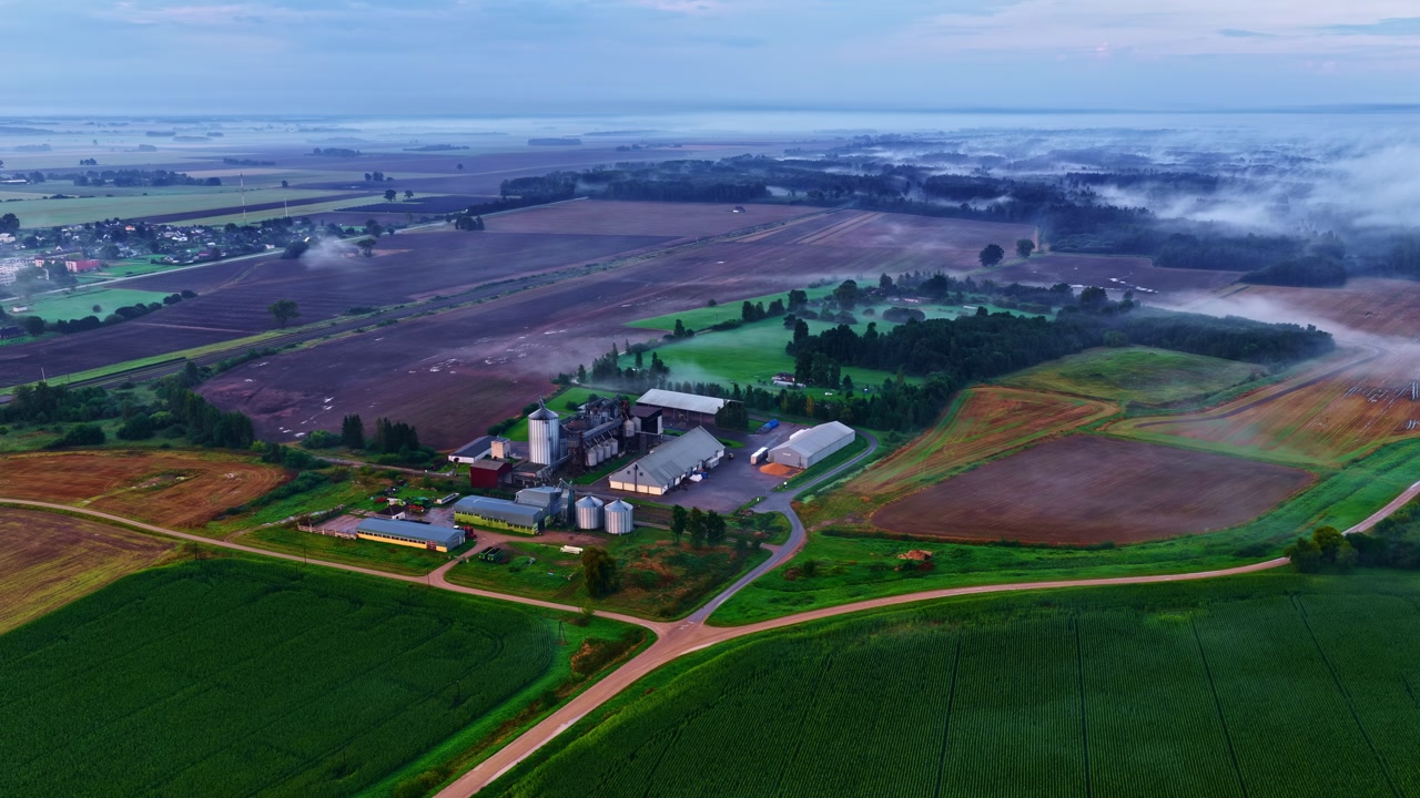Aerial pan shot of an industry located at rural area with green agricultural fields surrounding it.