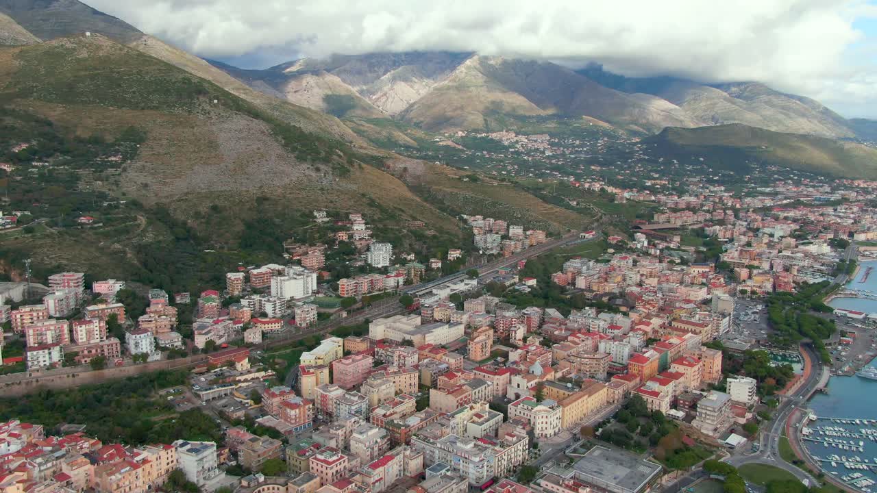 Flight above coastal downtown city buildings with view of seaport marina toward majestic glowing mountain range in background, Italy, aerial approach tilt up