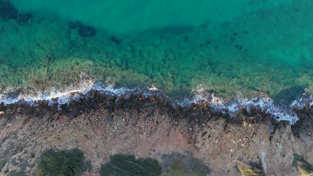 Aerial view of rocky coastline with crashing waves and clear turquoise waters in Crete.