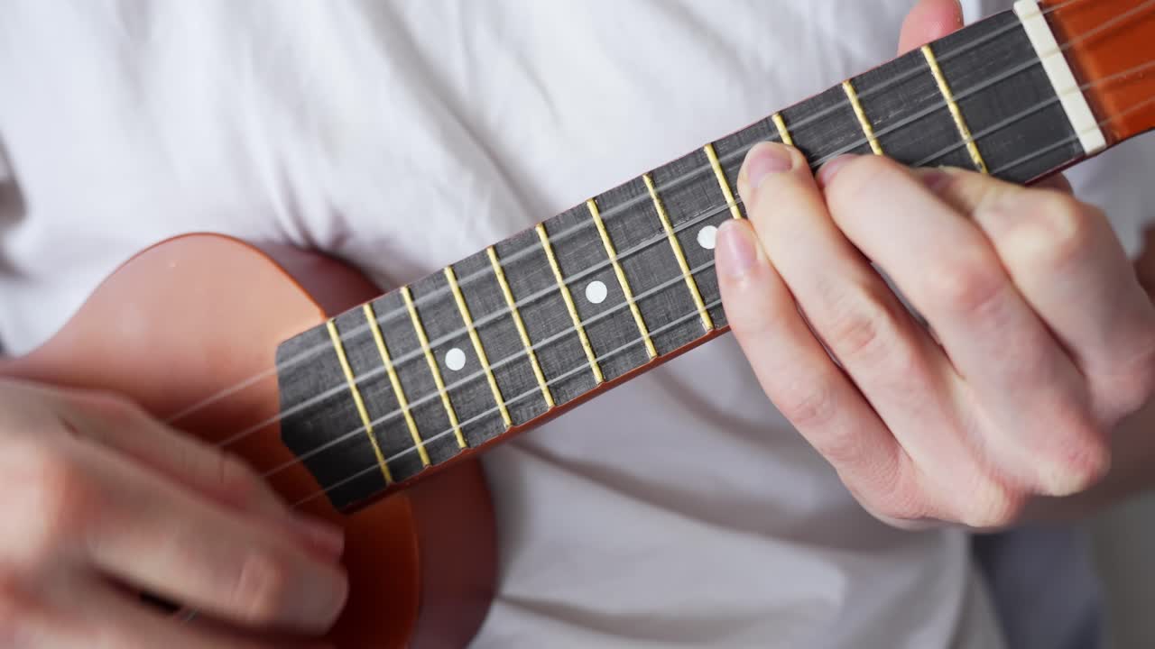 A close-up shot of a person playing a wooden ukulele indoors with focus on the hand positioning over the fretboard, capturing a calm home setting with natural daylight and musical expression.