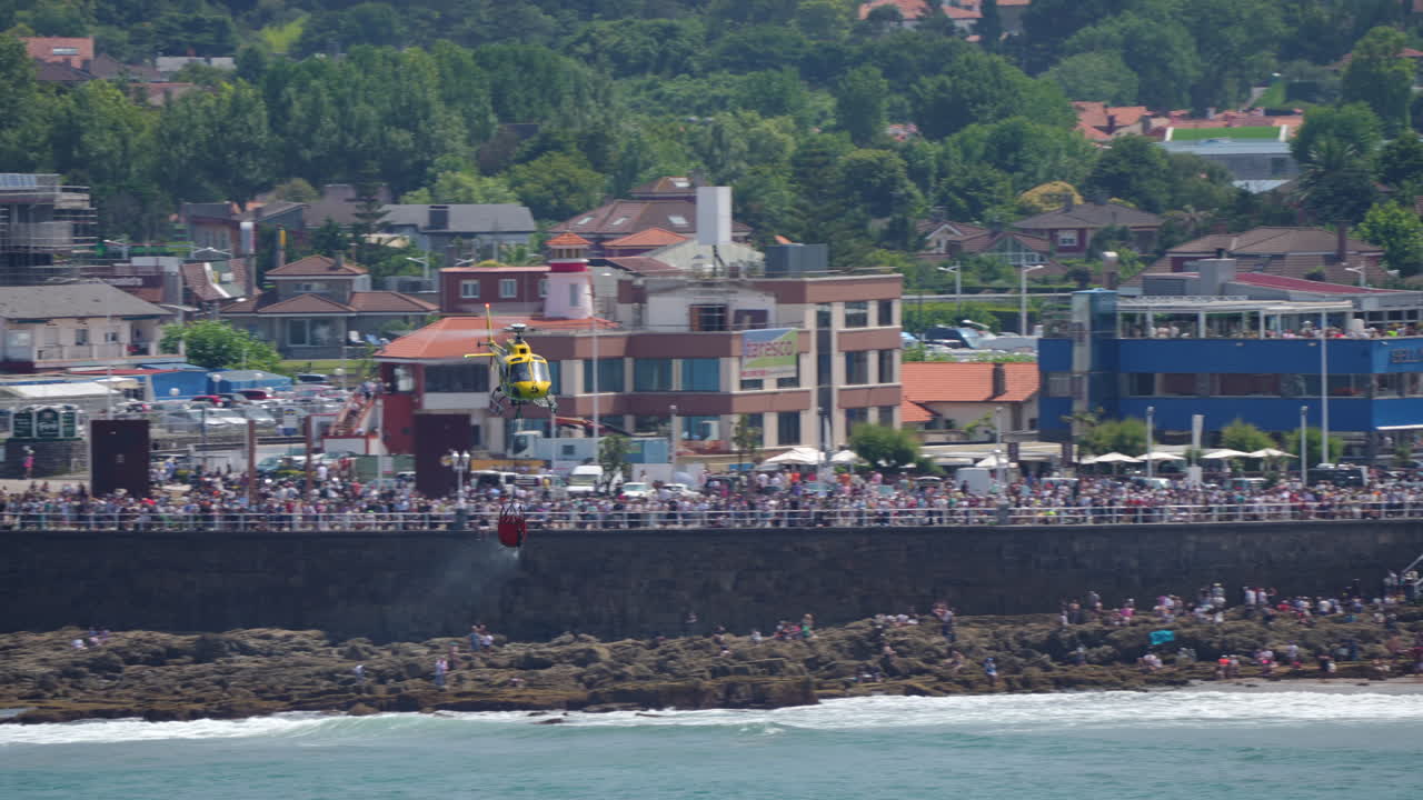 GIJON, SPAIN - JULY 27, 2025: Two firefighting helicopters release large volumes of water over the shoreline as part of a public demonstration during the Gijon airshow. Crowds gather along the promenade and beach to watch the display
