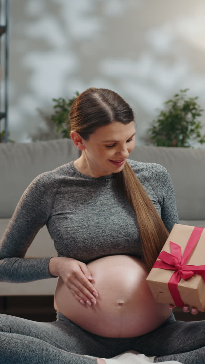 Expecting mother cradles her belly while holding a small gift box tied with a red ribbon indoors