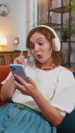 happy overjoyed young woman in wireless headphones using smartphone relaxing sitting on sofa at home