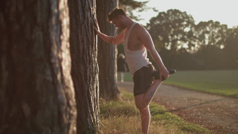 man in park stretching out his legs before he goes for a run whilst sun sets behind him