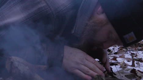 a hiker bends over the beginnings of a campfire from dried leaves and sticks as he uses his hands and face to shield the fire from the wind as the smoke begins to cloud around