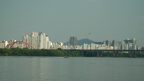 Persona-Irreconocible-Se-Cae-Mientras-Esquí-Acuático-En-El-Río-Hangang-Tirado-Por-Una-Vista-Lejana-De-Lancha-Rápida,-Tráfico-De-Automóviles-En-El-Puente-Seogang,-Edificios-De-Apartamentos-Y-Torre-Namsan-De-Seúl-Detrás-De-La-Isla-Bamseom