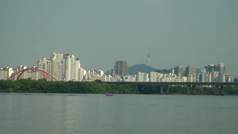 persona irreconocible se cae mientras esquí acuático en el río hangang tirado por una vista lejana de lancha rápida, tráfico de automóviles en el puente seogang, edificios de apartamentos y torre namsan de seúl detrás de la isla bamseom