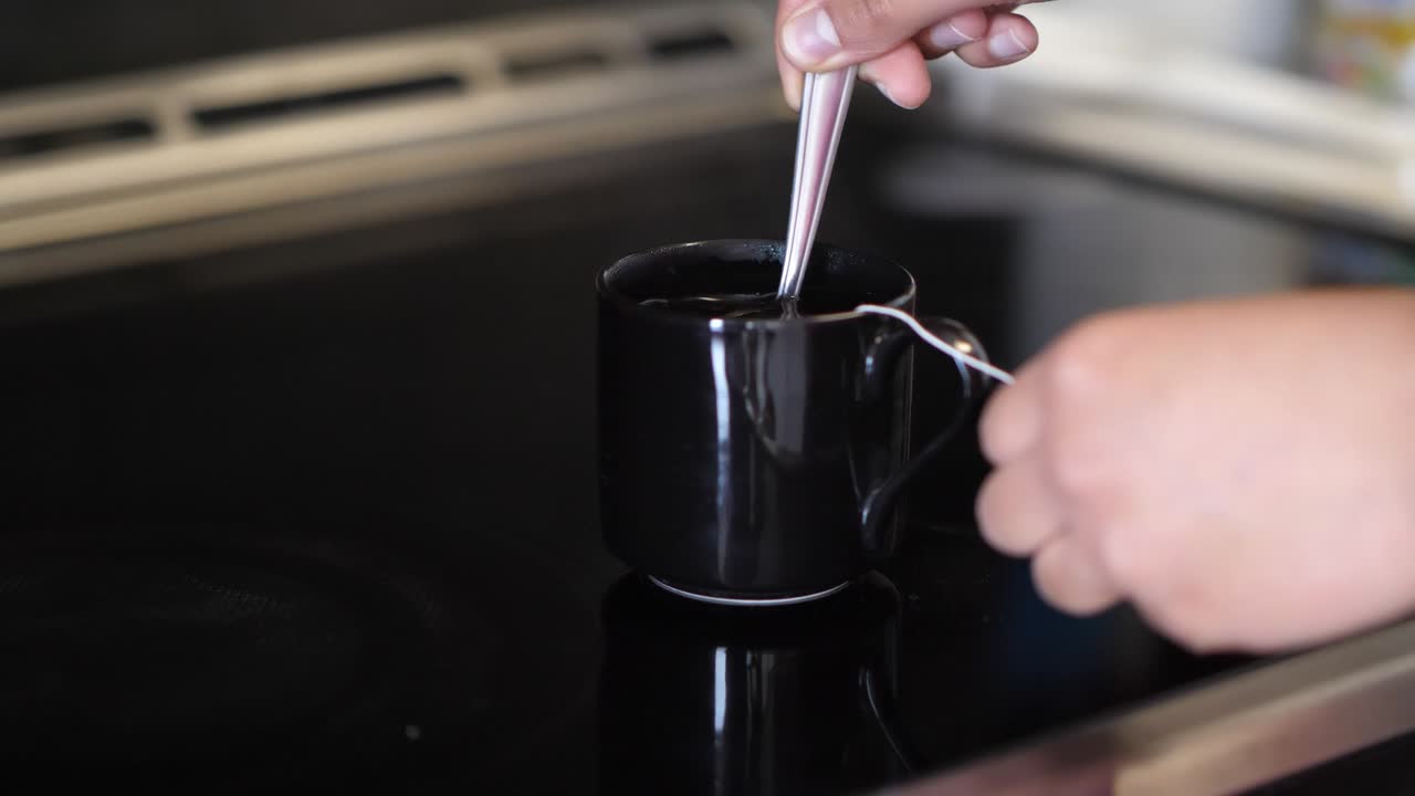 혼혈 아프리카계 미국인 여성의 손과 검은 차 컵의 클로즈업으로, 그녀는 차 봉투 을 들고 스토브에 앉아있는 는 뜨거운 물을 부드럽게 습니다.
