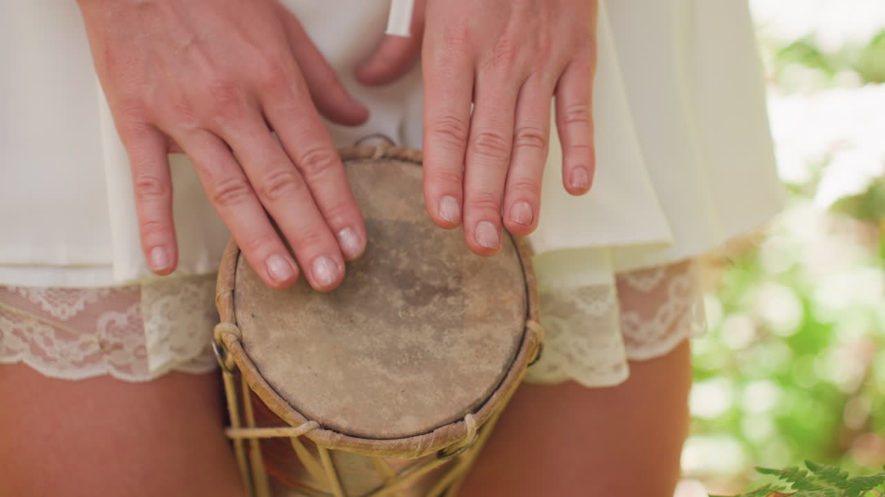 Close up woman in short white lace gown rhythmically playing talking drum outdoors, gentle sunlight highlighting natural movement of hands as she creates soothing tribal beat