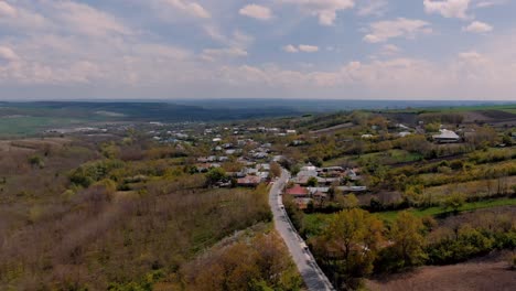 Aerial-view-over-an-old-village-road-and-houses,-agricultural-fields,-surrounding-hills,-partly-cloudy