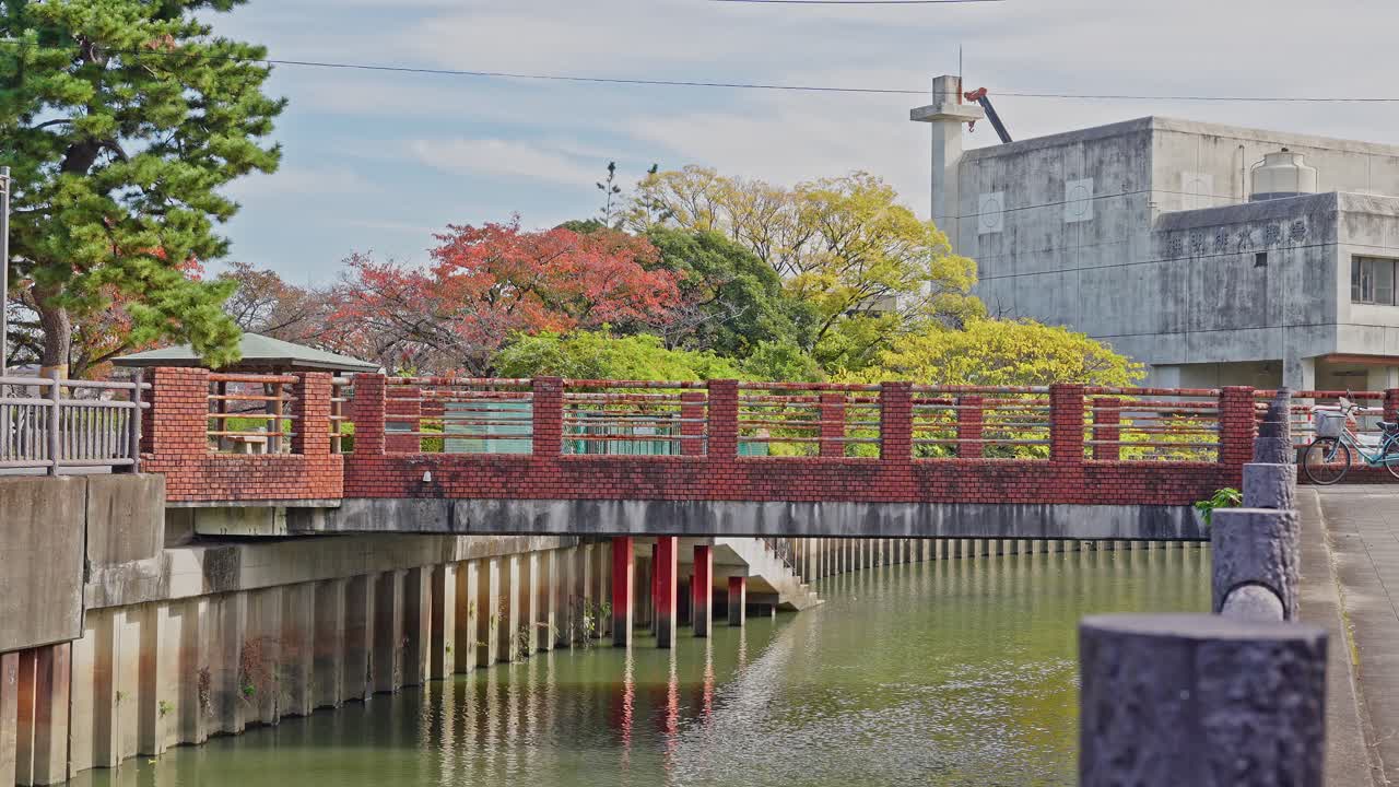 A small, rustic red pedestrian bridge crosses the river next to a modern building, framed by autumn trees