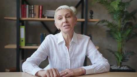 confident elderly woman teacher in white shirt making video call, online talking to webcam at classroom