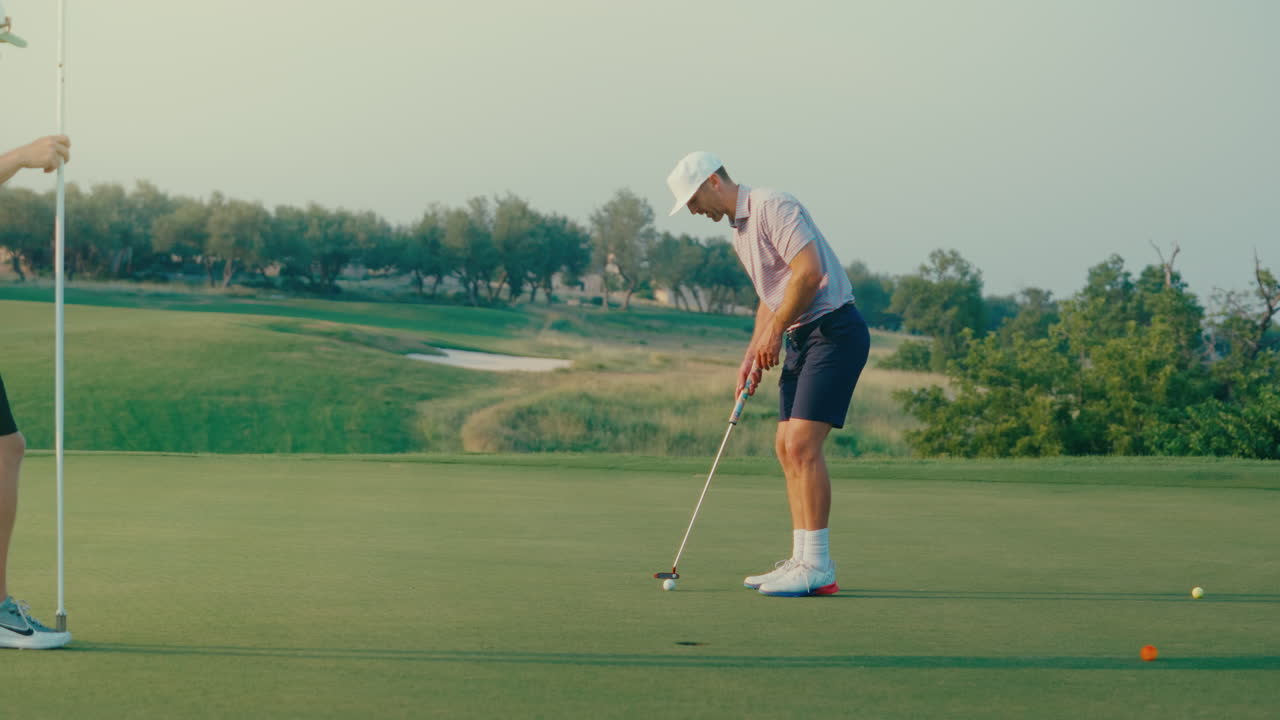 Male golfer reads the green and approaches his ball to putt while his playing partner pulls the flagstick and waits beside the hole. A quiet, focused moment showing teamwork and rhythm on the green.