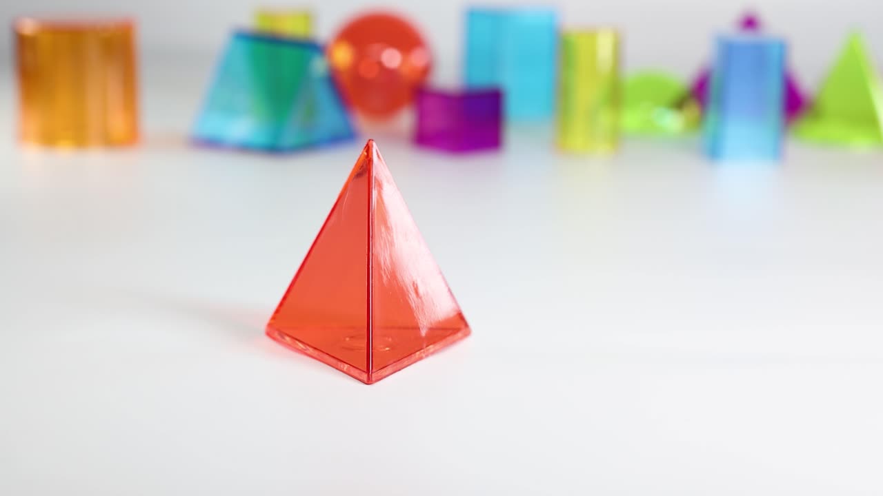 A hand interacts with a red plastic pyramid among colorful geometric shapes on a white table, under bright lighting