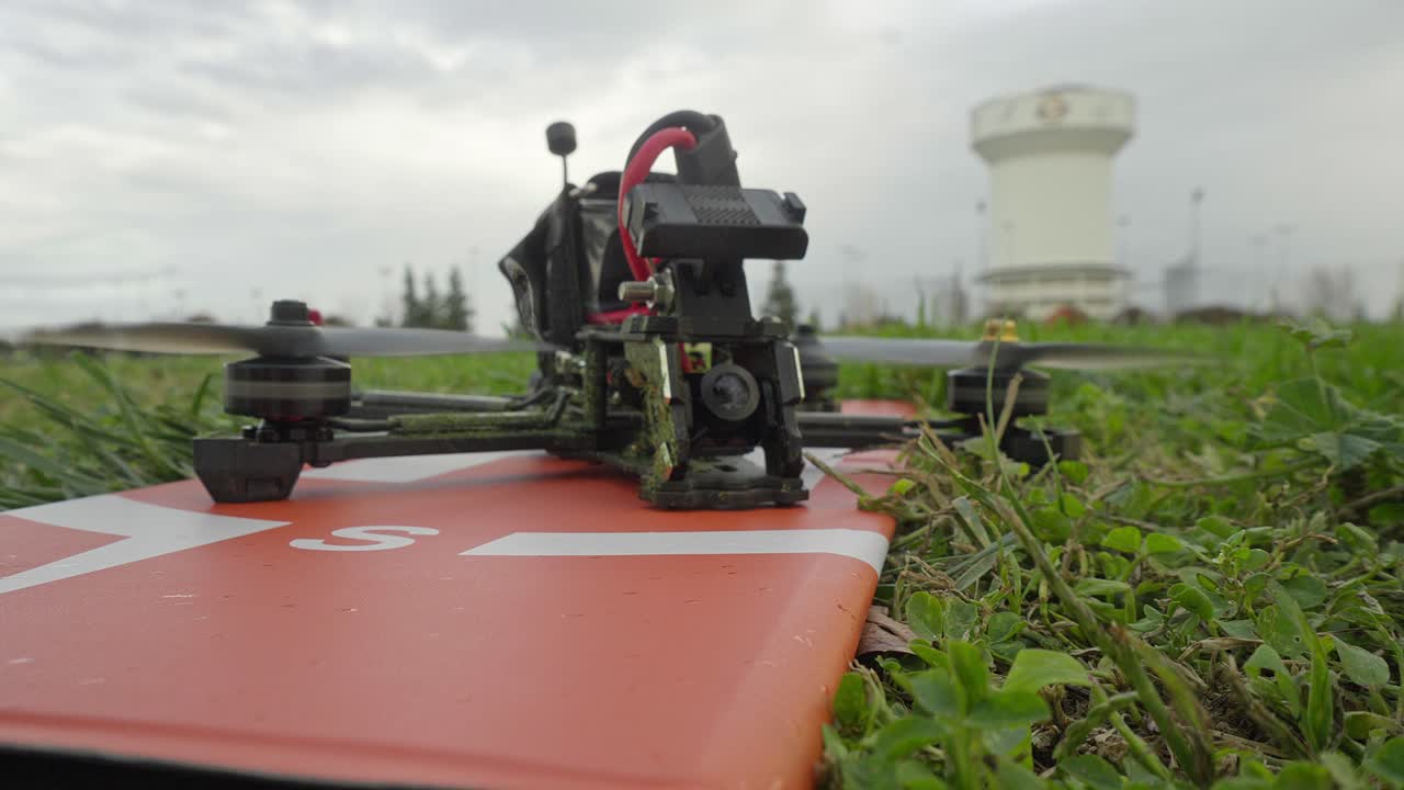 The drone sits among blades of grass while the arm switch is engaged, its motors gaining subtle momentum before launch