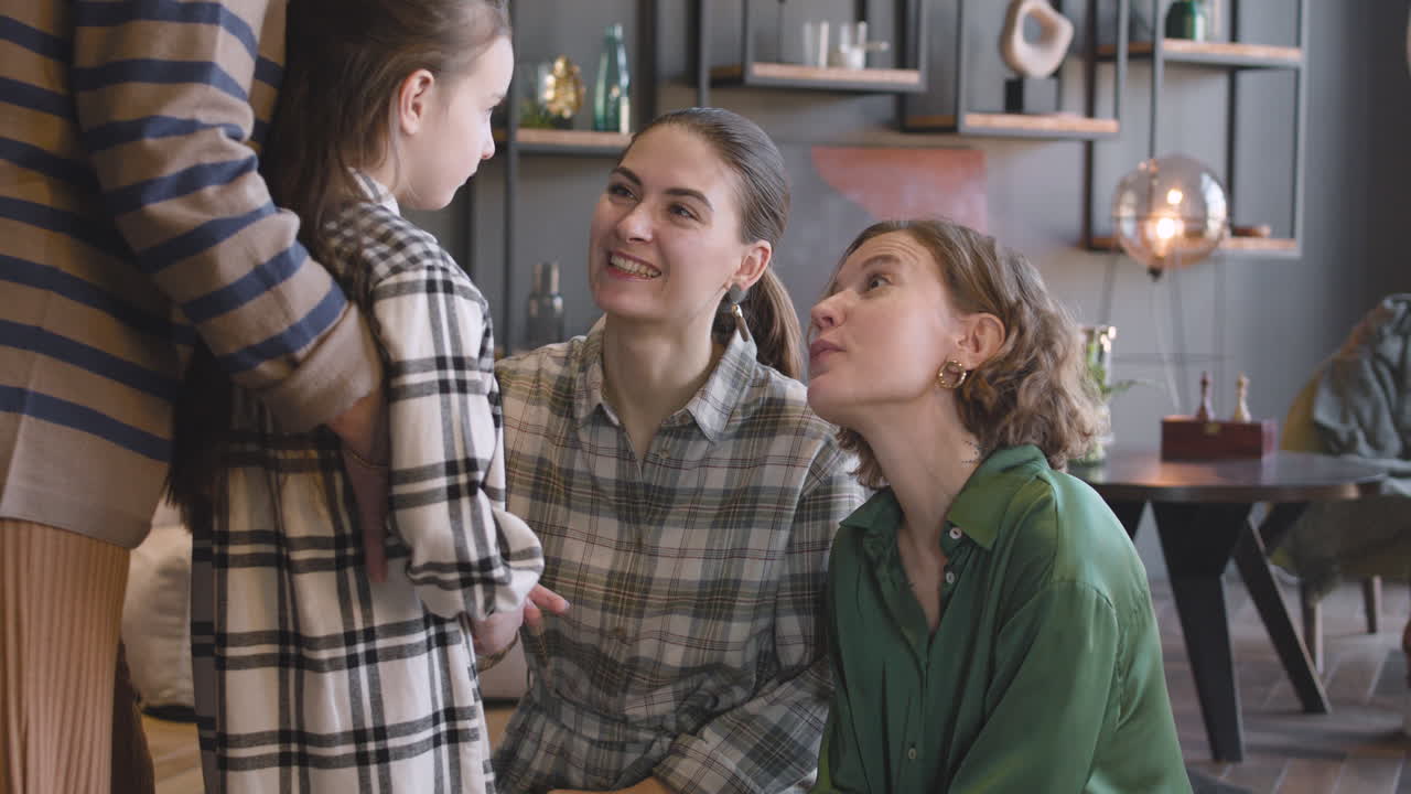 집에서 가족 모임을 하는 동안 알아볼 수 없는 할머니가 그녀를 뒤에서 껴안고 있는 동안 어린 소녀가 어머니와 이모와 이야기를 나누고 있습니다.