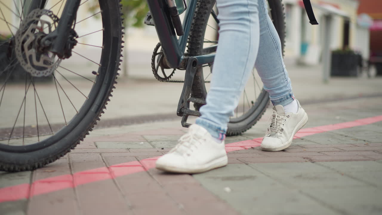 seorang wanita berdiri di samping sepeda di trotoar, close-up kaki, sepatu kets putih dan celana jin terang di dekat roda dan pedal sepeda, garis tepi jalan berwarna merah dan trotoar bertekstur, suasana jalanan kota, komuter kasual