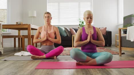 Happy-diverse-female-couple-doing-yoga-together-in-living-room,-meditating