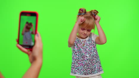 Cheerful-funny-little-teen-girl-kid-dancing-at-camera-filming-video-using-phone-on-chroma-key-wall