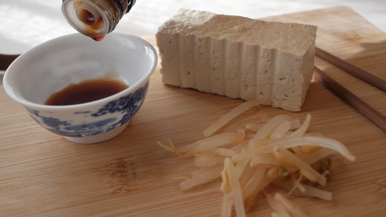 Tofu block and bean sprouts with soy sauce being poured into a small ceramic bowl