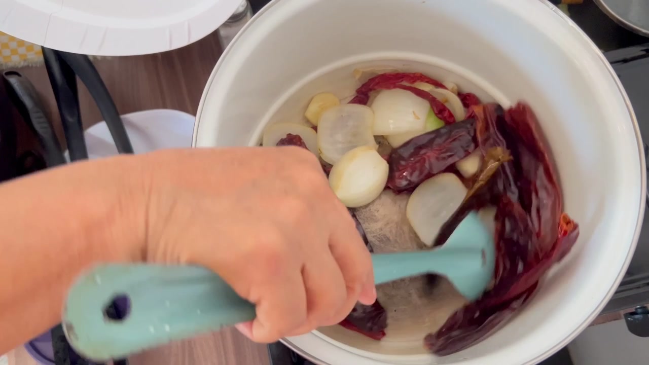 Eine Hand rührt eine Mischung aus getrockneten Chilis und gehackten Zwiebeln, frische Zutaten in einem Topf. Die Szene zeigt eine heimische Küche, in der traditionelle Kochtechniken hervorgehoben werden.