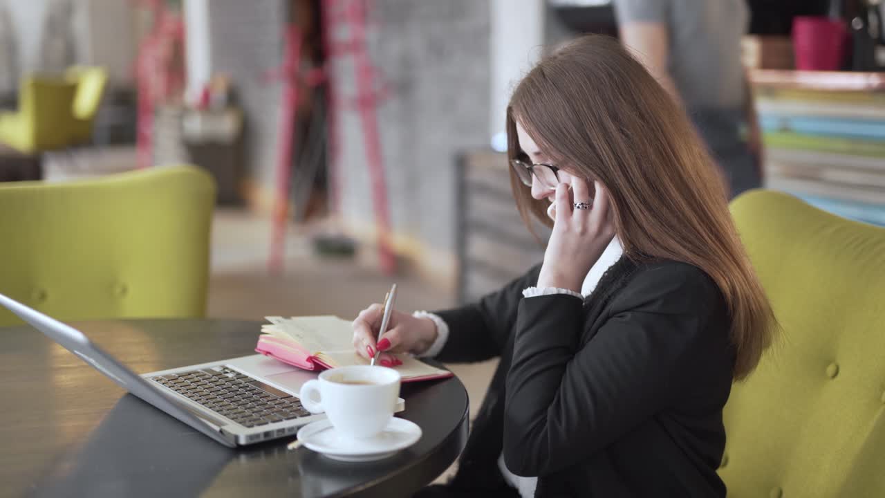 Donna che prende appunti al telefono davanti al laptop in una caffetteria